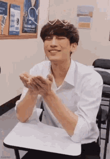 a man in a white shirt is sitting at a desk in a classroom with his hands folded and smiling .