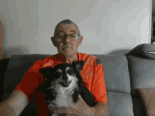 a man sitting on a couch holding a small black and white dog wearing an orange shirt that says ' adidas '