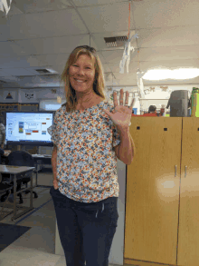 a woman wearing a floral shirt is waving in front of a classroom