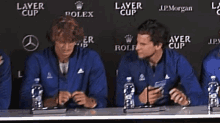 a group of men are sitting at a table with bottles of water in front of a wall that says layer cup