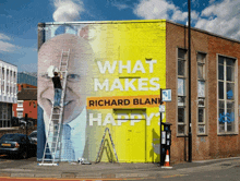 a man on a ladder paints a mural of richard blank on a yellow building