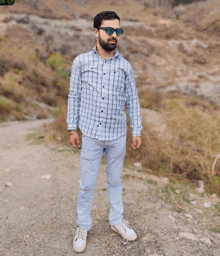 a man wearing sunglasses and a plaid shirt stands on a gravel road