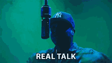 a man wearing a ny hat stands in front of a microphone with the words real talk written below him