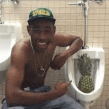 a shirtless man is sitting next to a urinal with a pineapple in it .