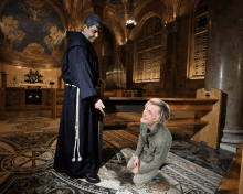 a man in a green jacket kneels in front of a priest