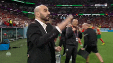 a man in a suit stands on a soccer field in front of a scoreboard that says live on it