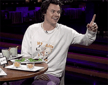 harry styles is sitting at a table with plates of food and a pearl necklace .