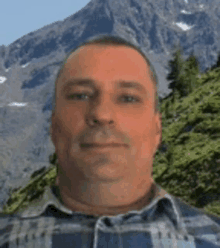 a man in a plaid shirt is standing in front of a mountain with trees in the foreground .