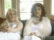 two women are sitting on a couch and one is wearing a white fur coat