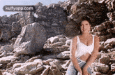 a woman in a white tank top is sitting on a rocky beach with her hair blowing in the wind .