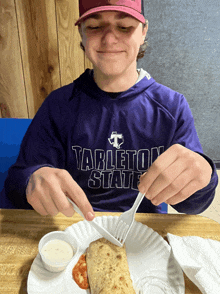 a boy wearing a purple tableton state shirt is cutting a pizza with a fork