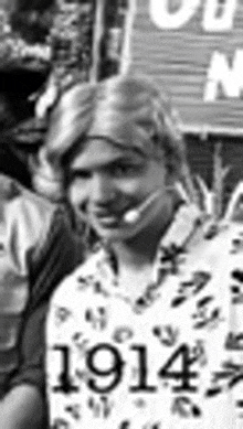 a black and white photo of a woman wearing a wig and a 1914 shirt .