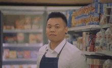 a man in a white shirt and blue apron stands in front of a shelf with a can of chicken noodle soup on it