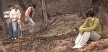 a group of people are standing and sitting in the woods . one of the people is sitting on the ground .