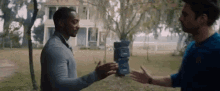 two men are standing next to each other in front of a house . one of the men is holding a stack of boxes .