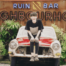 a young man sits on the front of a car in front of a sign that says ruin bar ghbourhc