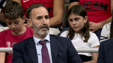 a man in a suit and tie sits in a crowd with a girl wearing a shirt that says cam