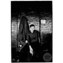 a black and white photo of a man smoking a cigarette while sitting in front of a brick wall .