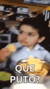 a young boy is sitting at a table eating chips with the words que puto written on the bottom .