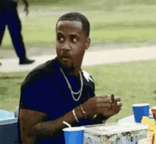 a man wearing a necklace is sitting at a table with a blue cup and a box of ice cream .