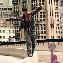 a man in a plaid shirt is standing on a ledge with his arms outstretched in front of a city skyline