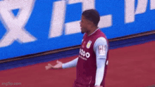 a soccer player in a maroon jersey is standing on a red carpet in front of a large blue sign .