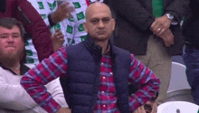 a man in a plaid shirt and vest is sitting in a crowd of people .