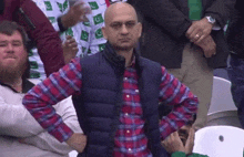 a bald man wearing a plaid shirt and a vest is sitting in a crowd of people .