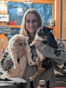 a woman holds two cats in front of a sign that says " the cook is not responsible for hair in your food "