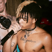 a shirtless man with dreadlocks is singing into a microphone while another man holds a camera behind him