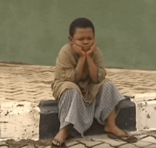 a young boy is sitting on a curb with his hands on his face and a sad look on his face .