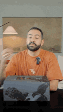 a man with a beard wearing an orange t-shirt is sitting at a table with a box in front of him .