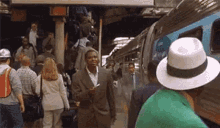 a man in a suit and hat is standing in front of a train at a train station talking on a cell phone .