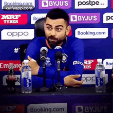 a man in a blue shirt is sitting at a table with bottles of water and talking into a microphone .