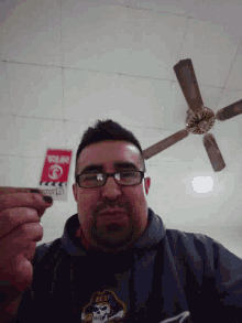 a man wearing glasses and a shirt with a skull on it is smoking a cigarette in front of a ceiling fan