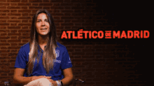 a woman in a blue shirt is sitting in front of a brick wall with the words atletico de madrid above her