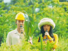 a man wearing a hard hat and a woman wearing a furry hat are singing into microphones in the woods .