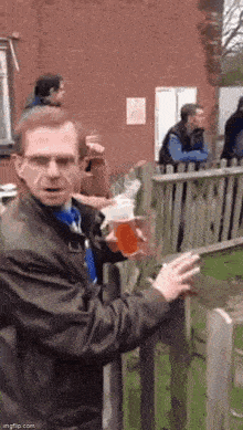 a man is holding a glass of beer and smoking a cigarette while standing next to a wooden fence .