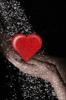 a hand is holding a red heart that is surrounded by sparkles