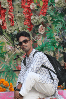 a man wearing sunglasses and a backpack is kneeling down in front of flowers