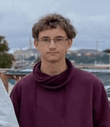 a young man wearing glasses and a purple sweatshirt is standing in front of a building .