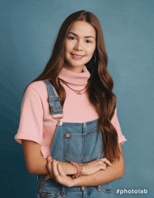 a woman wearing overalls and a pink turtleneck stands with her arms crossed in front of a blue background