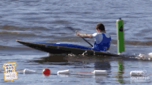 a man in a blue and white kayak is in the water near a green pole that says olympic channel