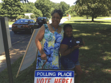 two women standing next to a polling place vote here sign