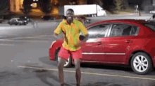 a man in a yellow and green shirt is dancing in front of a red car in a parking lot .