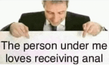 a man in a suit and tie is holding a sign that reads `` the person under me loves receiving anal '' .