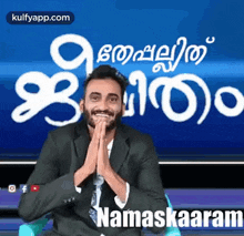 a man in a suit and tie is smiling with his hands folded in front of a screen that says namaskaaram