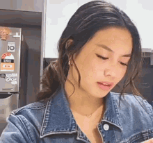 a woman wearing a denim jacket and a necklace is standing in front of a refrigerator in a kitchen .