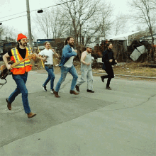 a man in an orange vest is playing a guitar while a group of men are running down the street