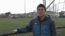 a man wearing a blue jacket with a yellow logo on the front stands on a fence in front of a soccer field